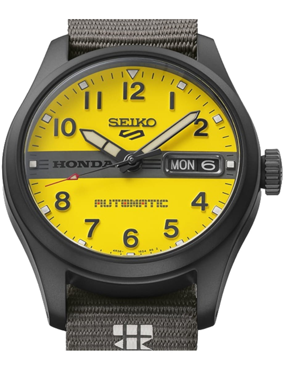 Yellow Seiko automatic watch with black case, gray fabric strap, and day/date display reading MON 6 on the right side of the dial.