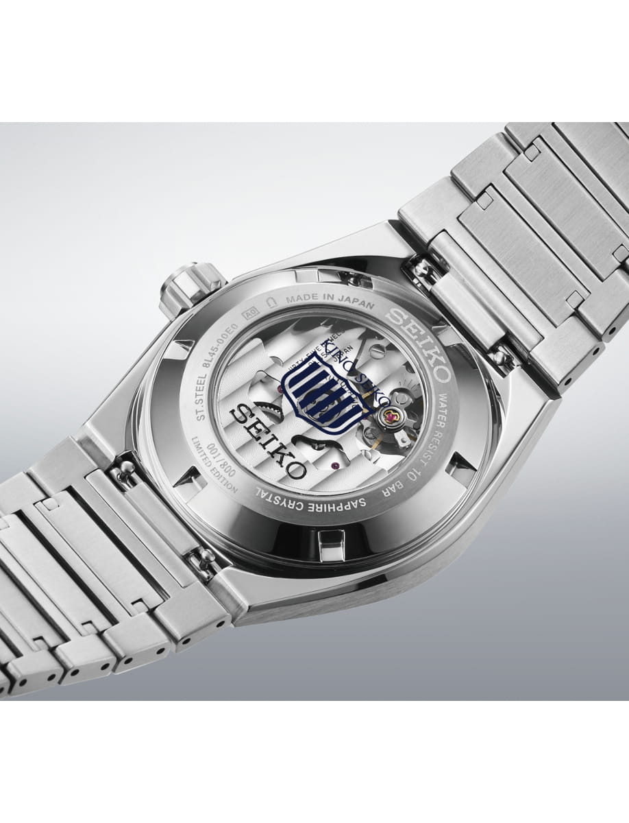 Back view of a stainless steel Seiko wristwatch with a transparent case revealing its mechanical movement and a blue emblem shield motif on the rotor.