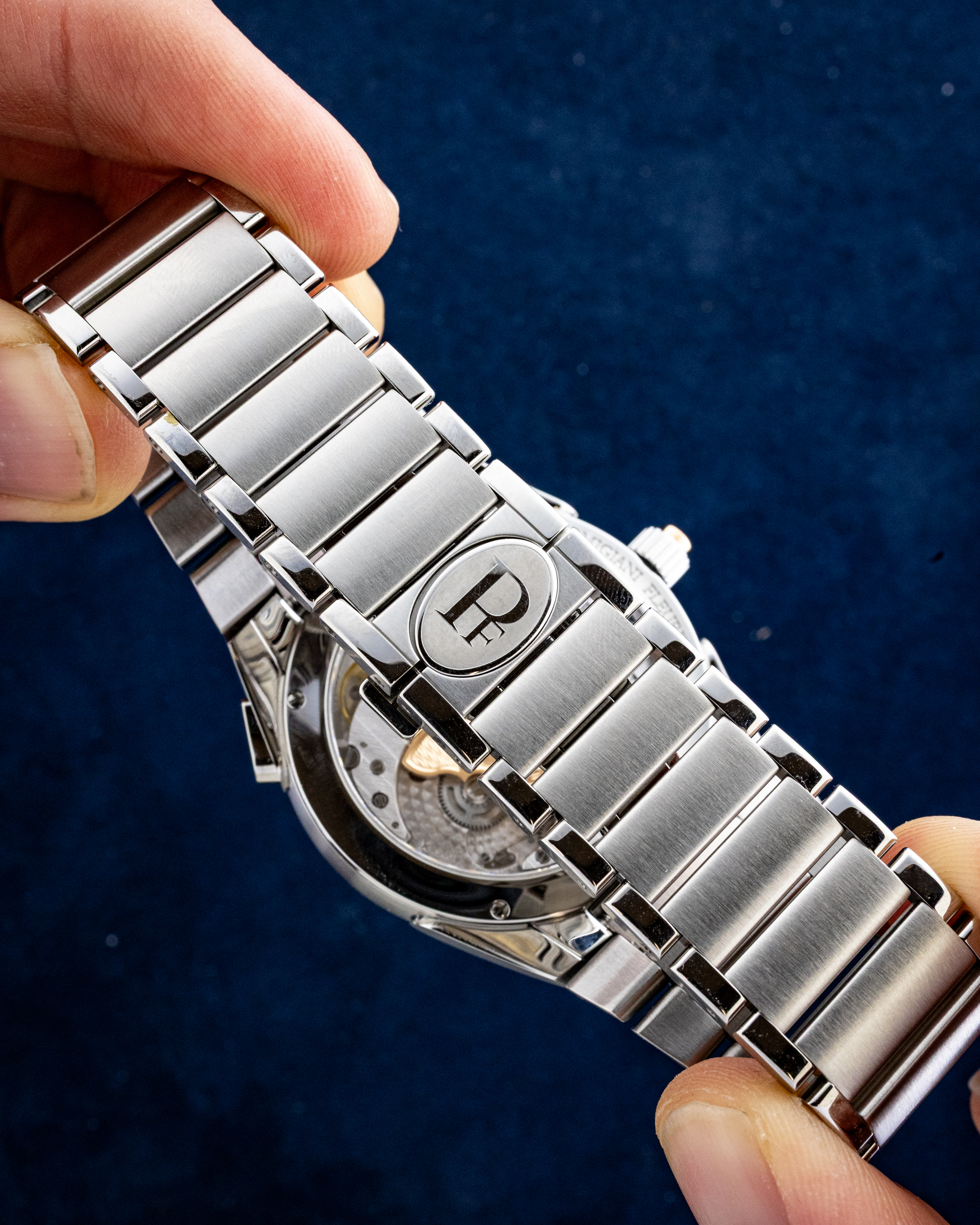 Close-up of a stainless steel watch bracelet being held by fingers, showing the clasp with a circular logo, against a dark blue background.
