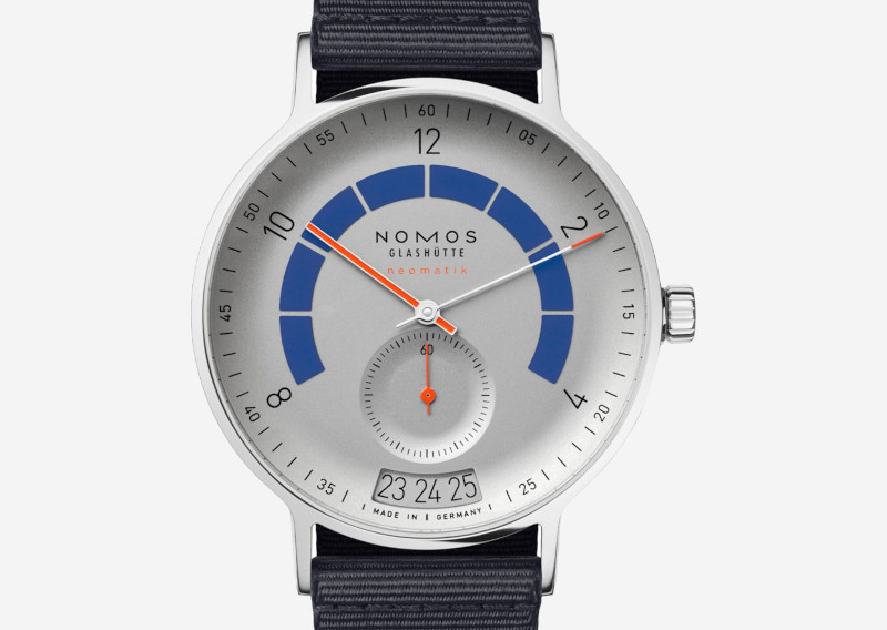 Nomos Glashütte watch with silver dial, blue power-reserve arc, orange hands, small seconds subdial, and date window at 6‑o'clock.