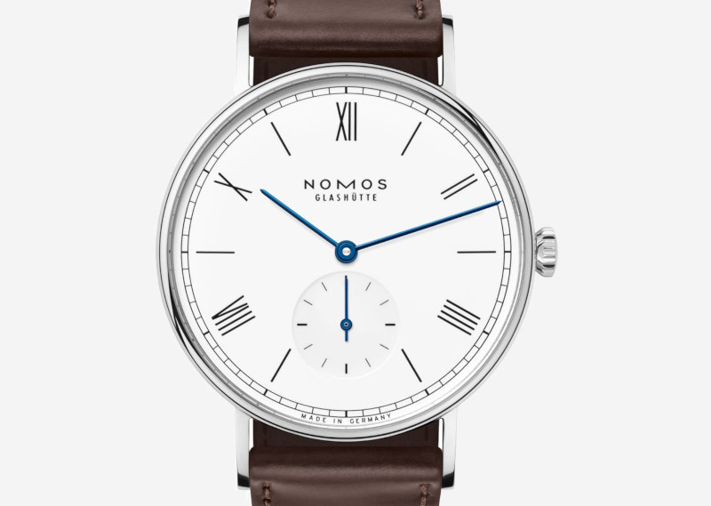 Nomos Glashütte wristwatch with white dial, blue hands, Roman numerals, brown leather strap.