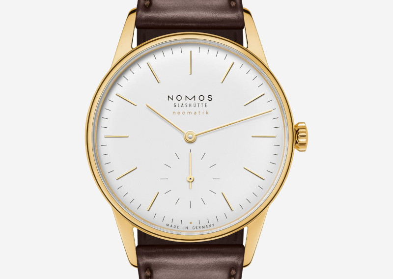 Gold-toned wristwatch with white dial, slim gold indices and hands, brown leather strap, NOMOS Glashütte Neomatik branding.
