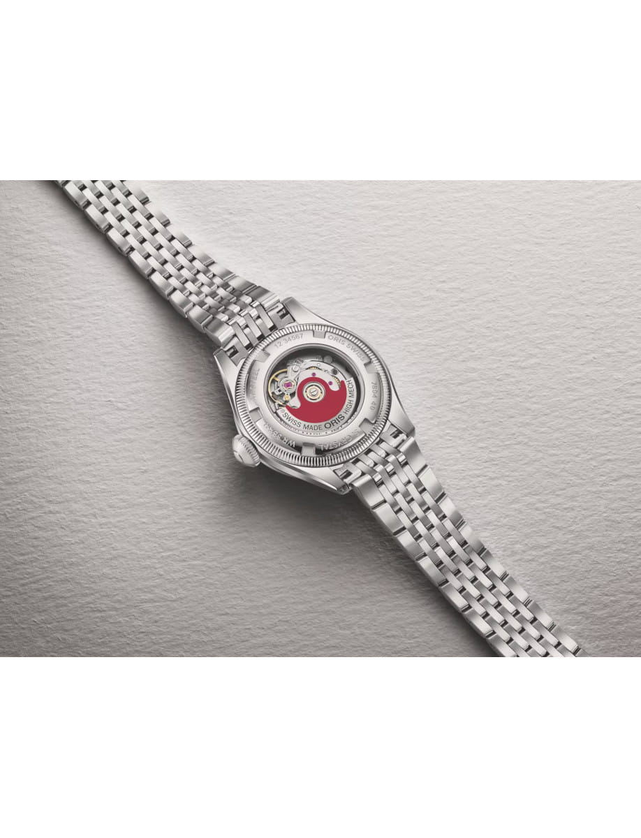 Stainless steel wristwatch with a transparent back showing the mechanical movement and red rotor, resting on a textured light surface with a metal bracelet edge visible.