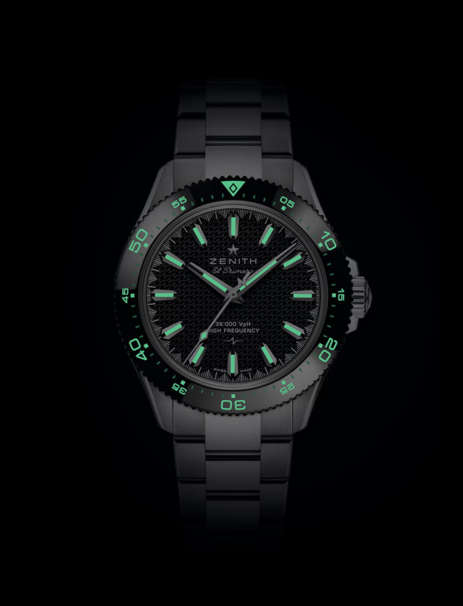 Close-up of a stainless steel Zenith wristwatch with glowing green lume on hands and hour markers against a black background. It has a date window at 3 o'clock and a green-lit bezel.