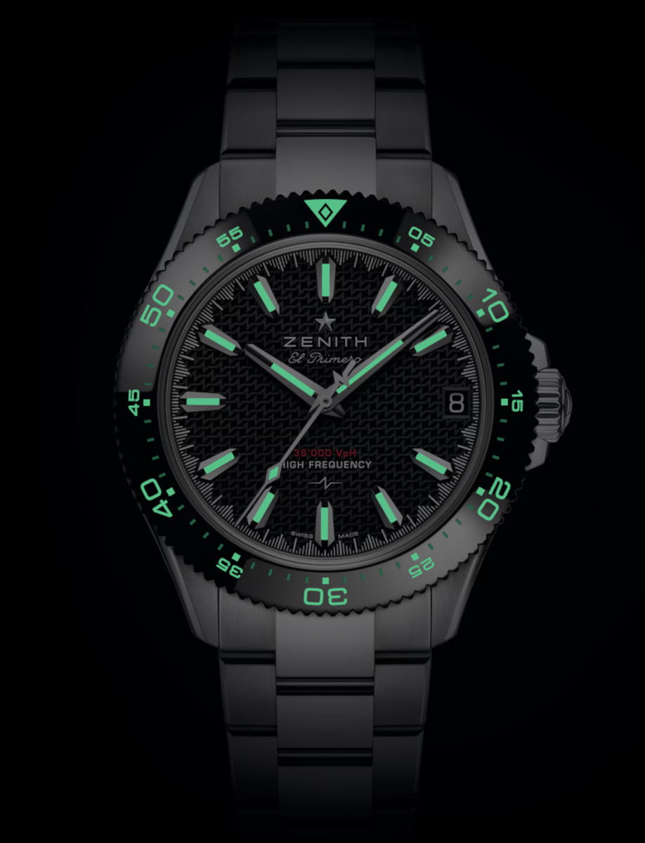 Stainless steel Zenith watch with glowing green hour markers and bezel numerals on a black dial, date window at 3 o'clock