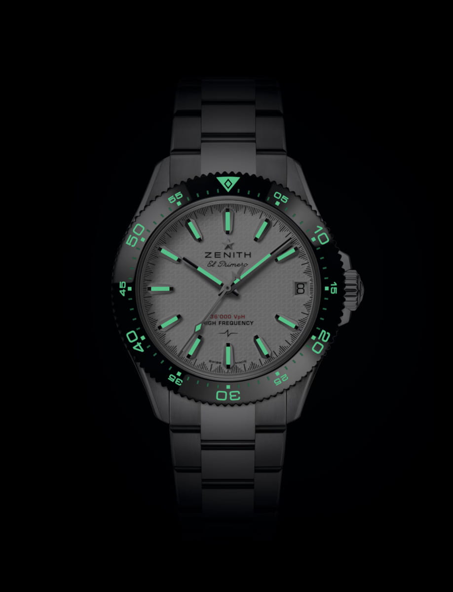 Close-up of a stainless steel Zenith wristwatch with glowing green lume on hands and hour markers against a black background. It has a date window at 3 o'clock and a green-lit bezel.