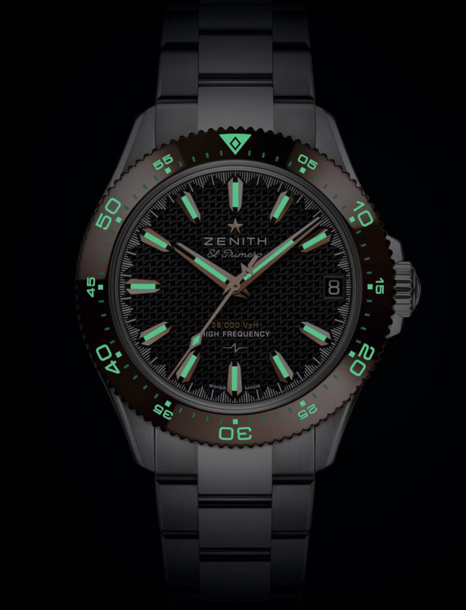 Zenith El Primero wristwatch with a black textured dial, glowing green luminescent markers and hands, date window at 3 o'clock, and a metal bracelet.