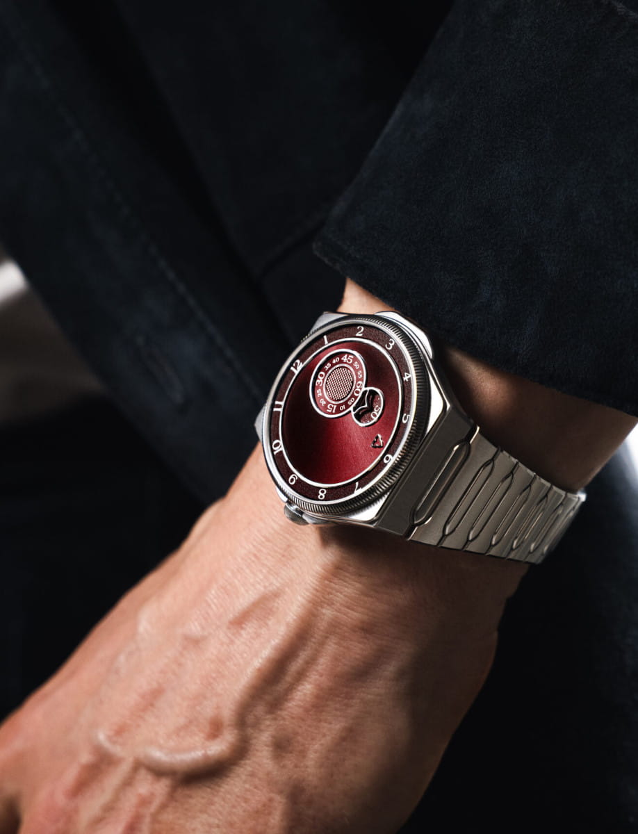 Close-up of a stainless steel wristwatch on a brown-skinned wrist, featuring a deep red circular dial and brushed band