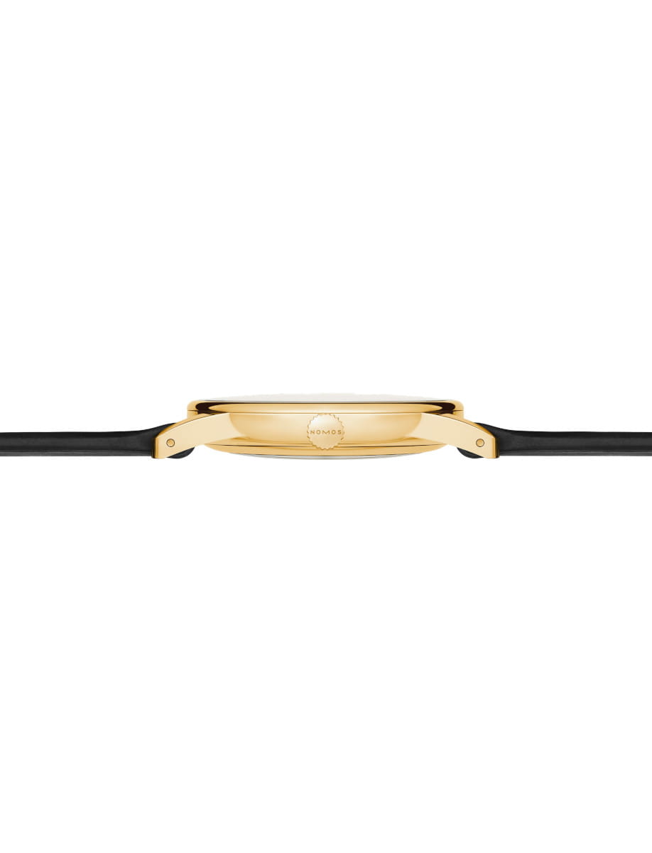 Side view of a gold-toned wristwatch with a black leather strap, showing the crown labeled NOMOS.