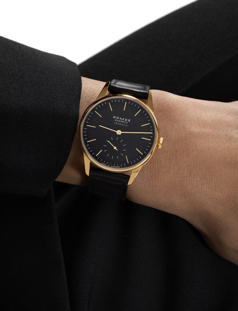 Elegant gold wristwatch with a black dial and leather strap on a person's wrist in a black sleeve.