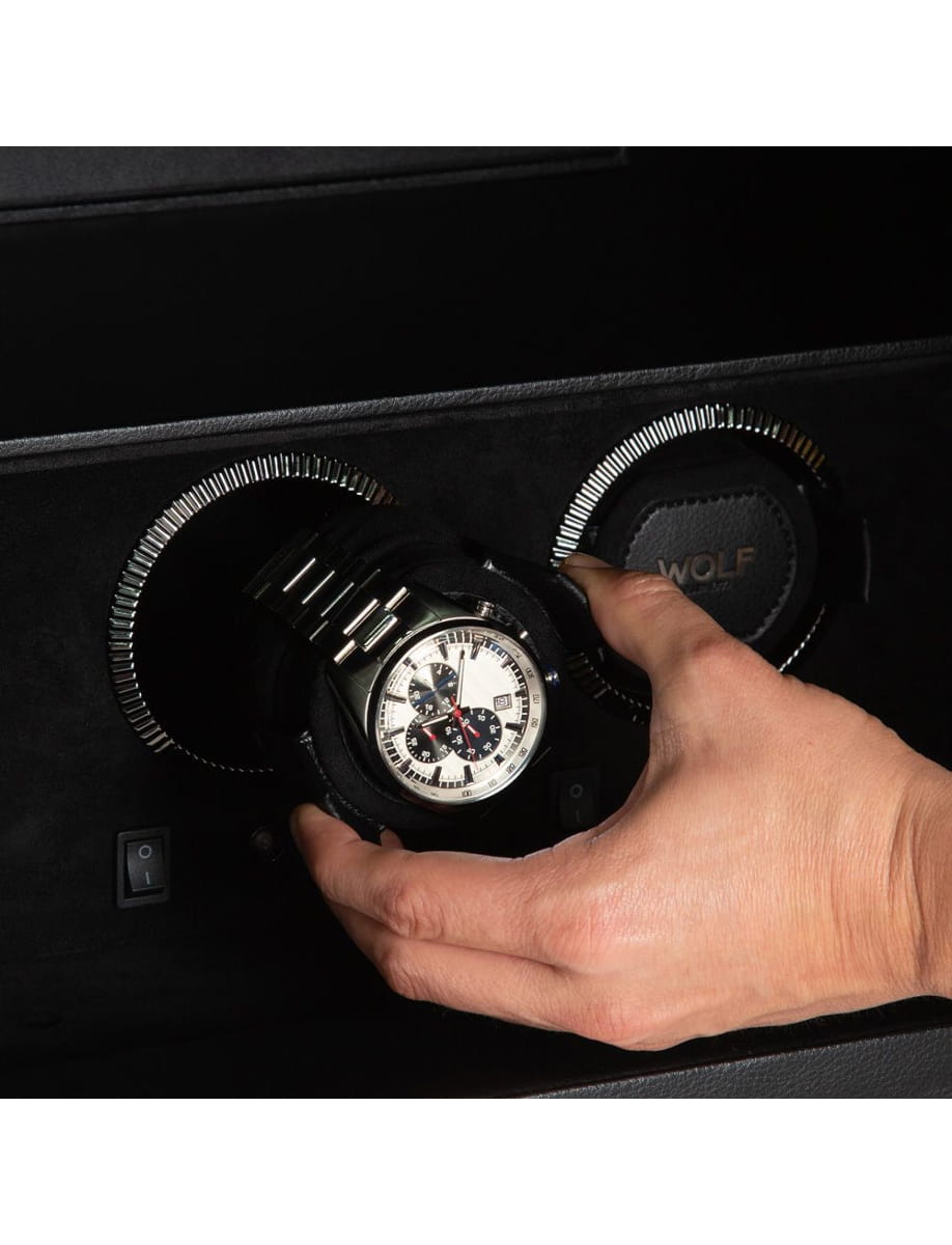 Person places a silver chronograph wristwatch into a safe vault slot, hand guiding it into the circular opening.
