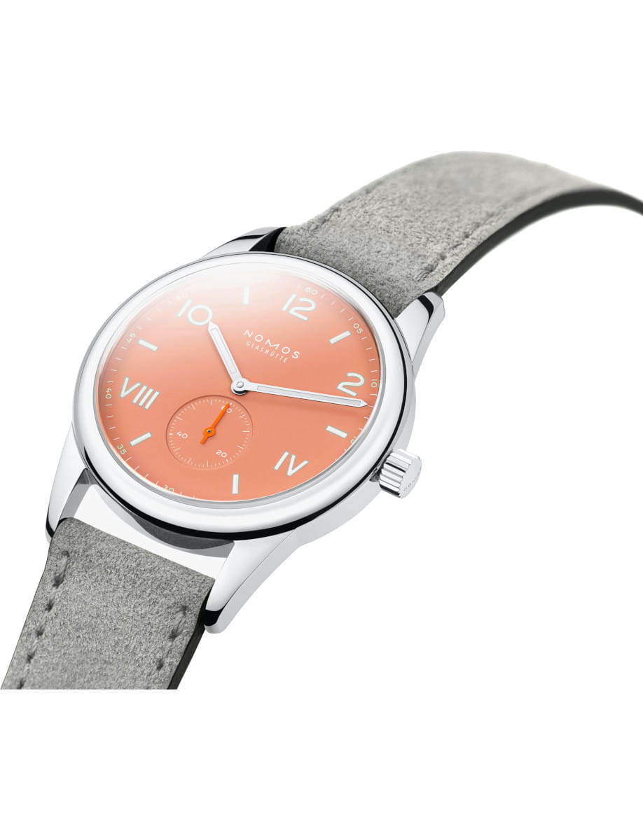Nomos Glashütte wristwatch with a peach dial, silver case, and gray suede strap viewed from an angled side.