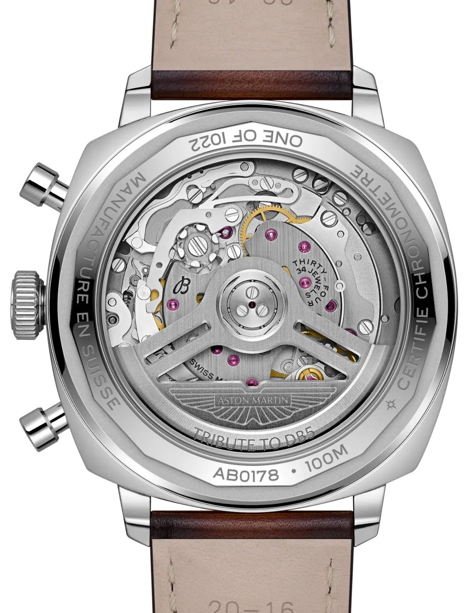 Exhibition caseback of a stainless steel Aston Martin watch, revealing the intricate mechanical movement with gears and jewels; engraved 'Tribute to DB5' and 'Aston Martin' visible, brown leather strap in view.