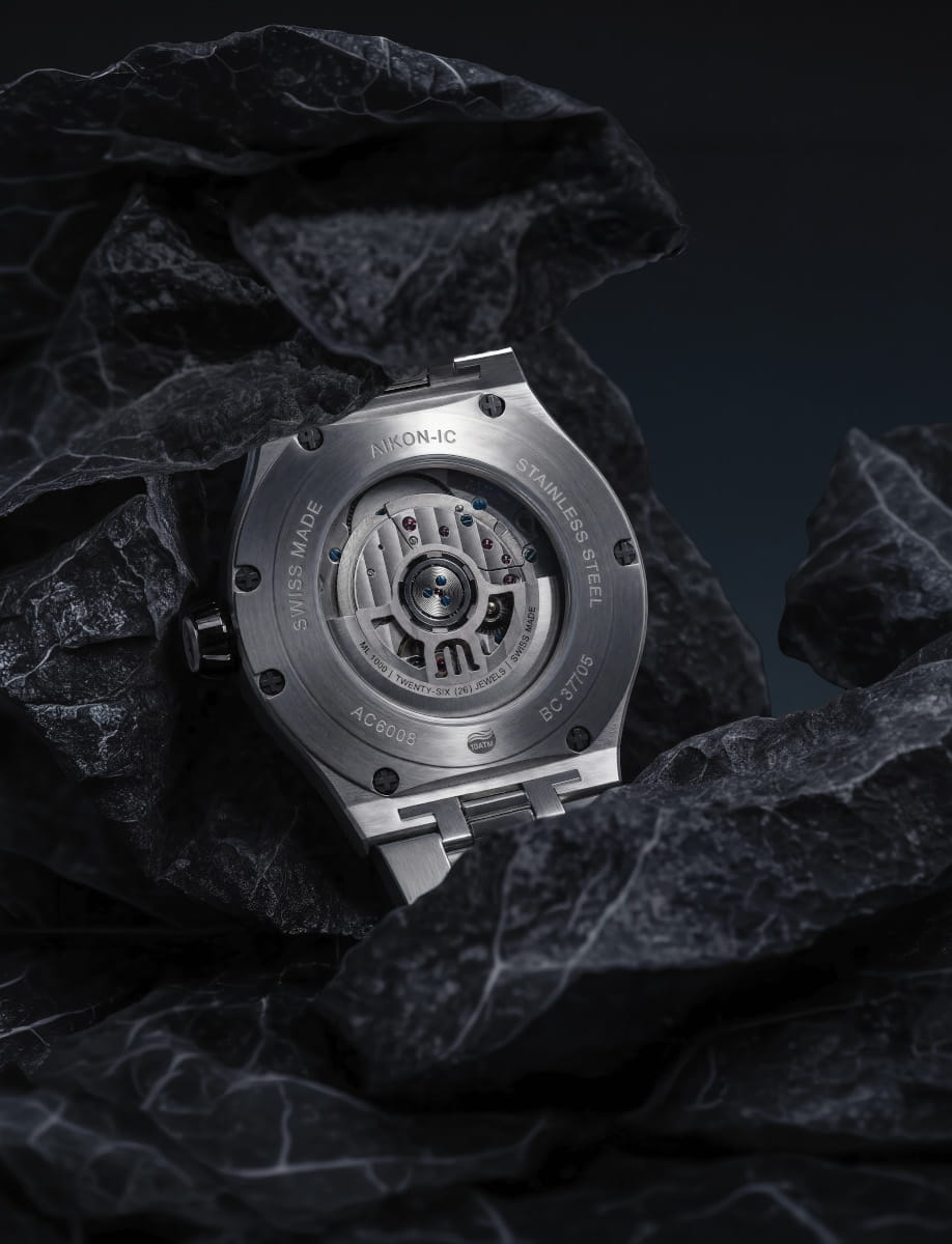 Back view of a Maurice Lacroix stainless steel wristwatch with a visible mechanical movement resting among dark rocks.