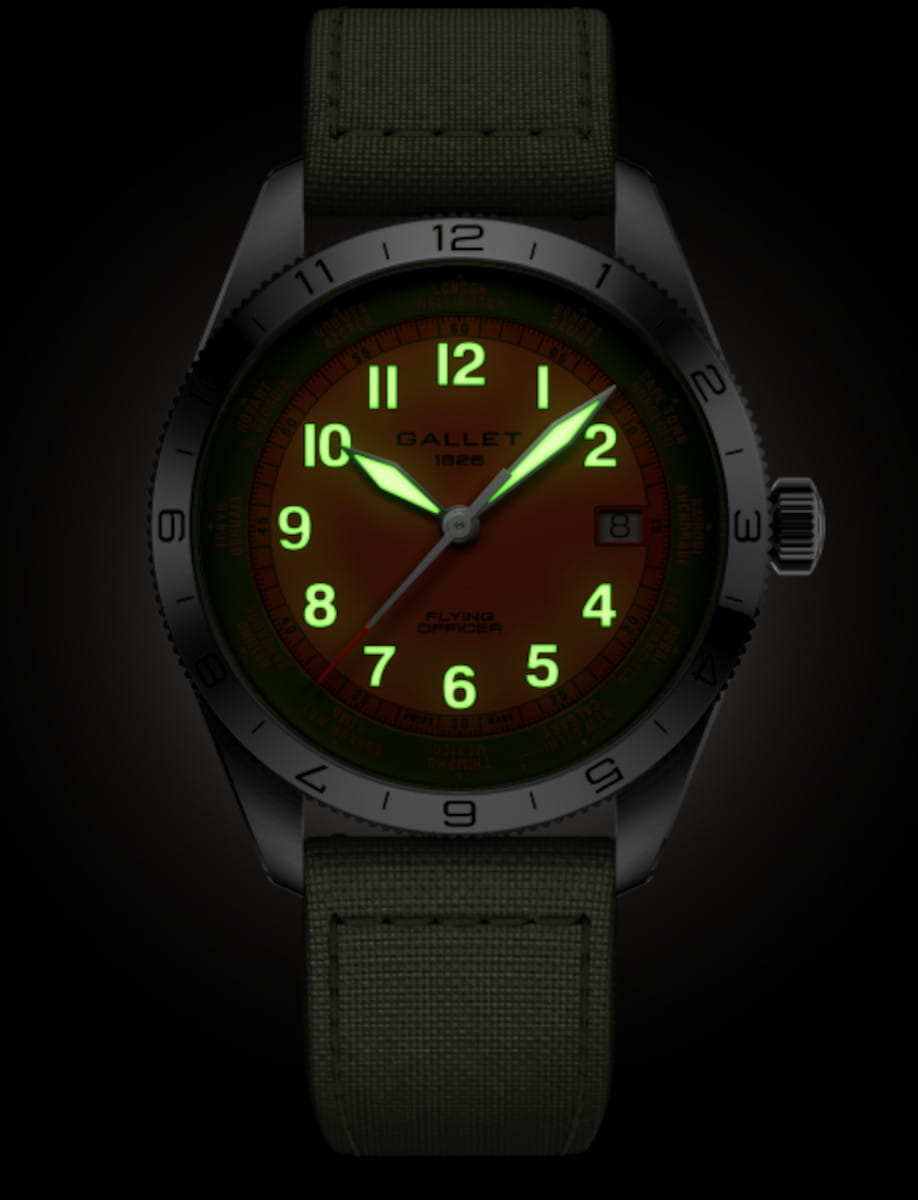 Gallet Flying Officer wristwatch with an orange dial, green luminescent numerals and hands, a green fabric strap, and a date window at 3 o'clock against a dark background.