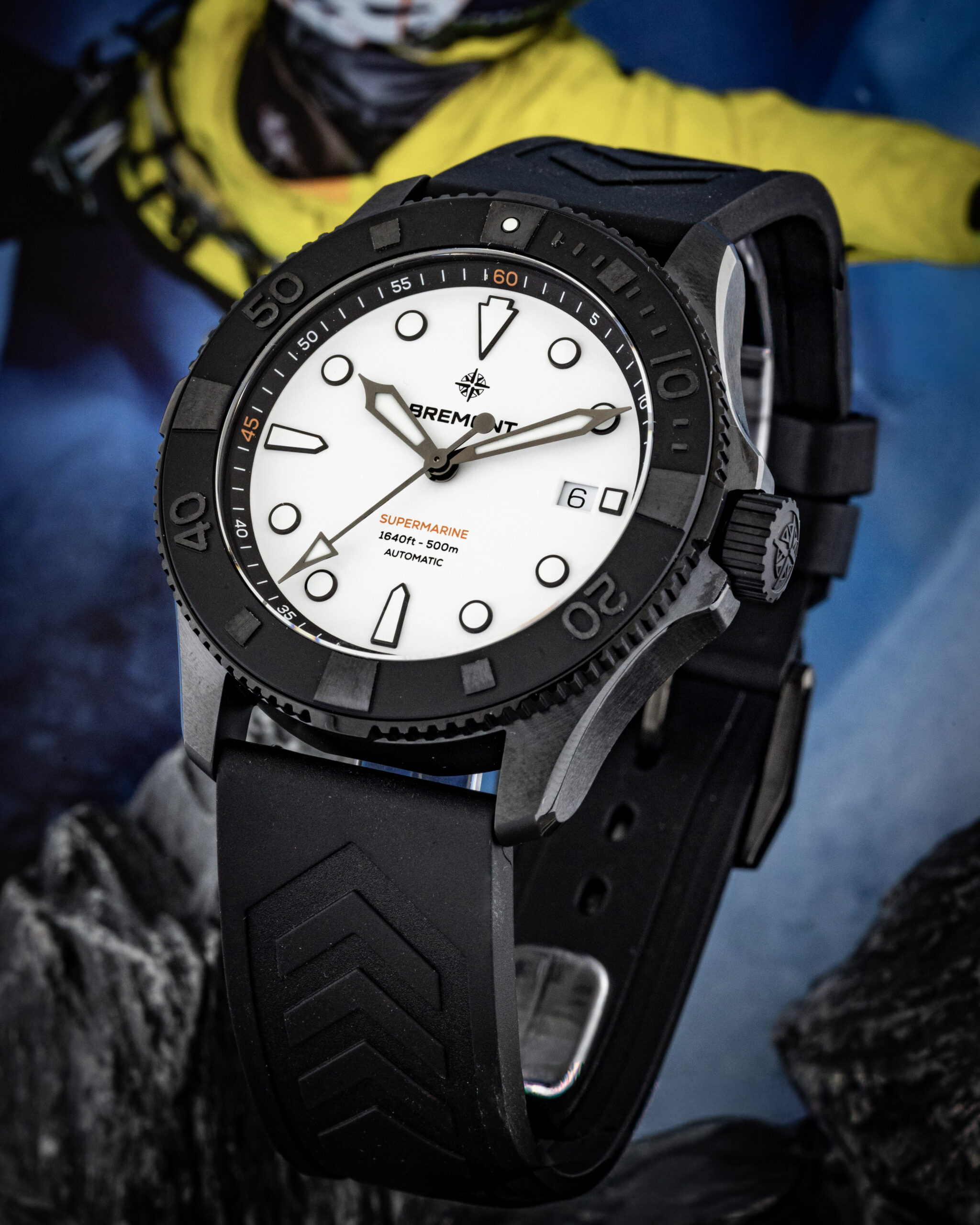 Close-up of a black Bremont dive watch with white dial on a rugged rock surface and a yellow jacket in the blurred background.
