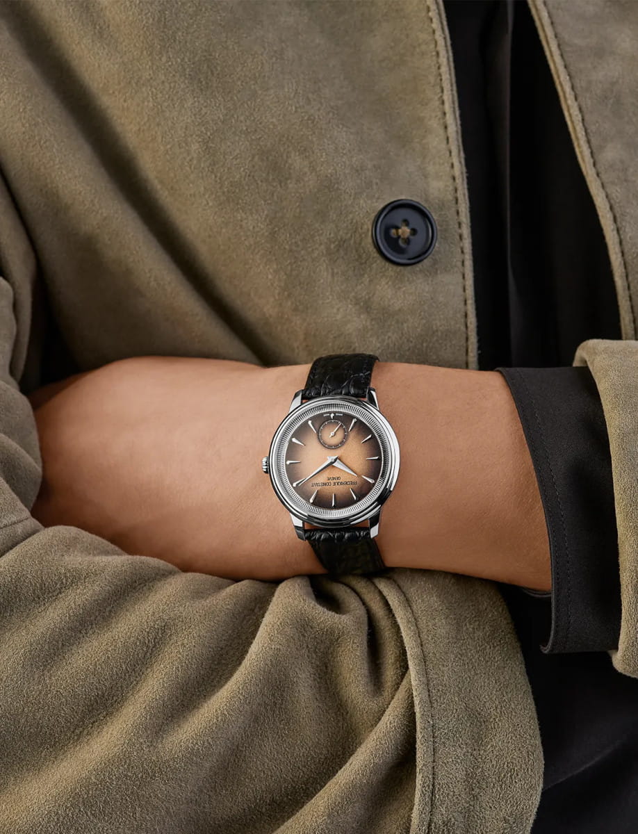 Wristwatch with a black to salmon gradient dial on a black leather strap worn on a forearm, against a beige suede coat.