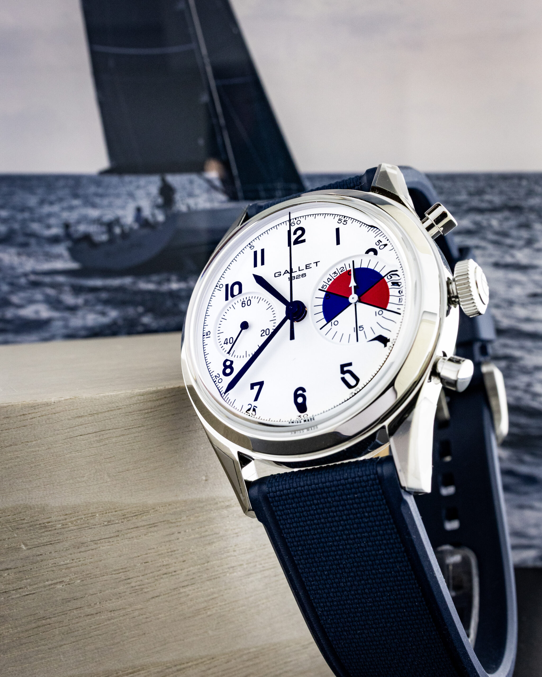 Close-up of a white analog chronograph watch with a blue strap resting on a wooden surface, with a blurred sailboat and sea in the background.