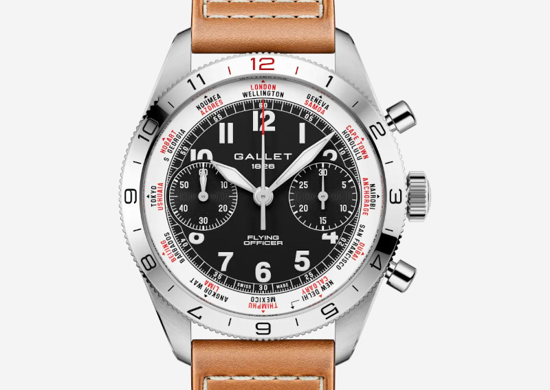 A stainless steel watch with black dial, white numerals, three subdials, red city names around the inner ring, and a tan leather strap