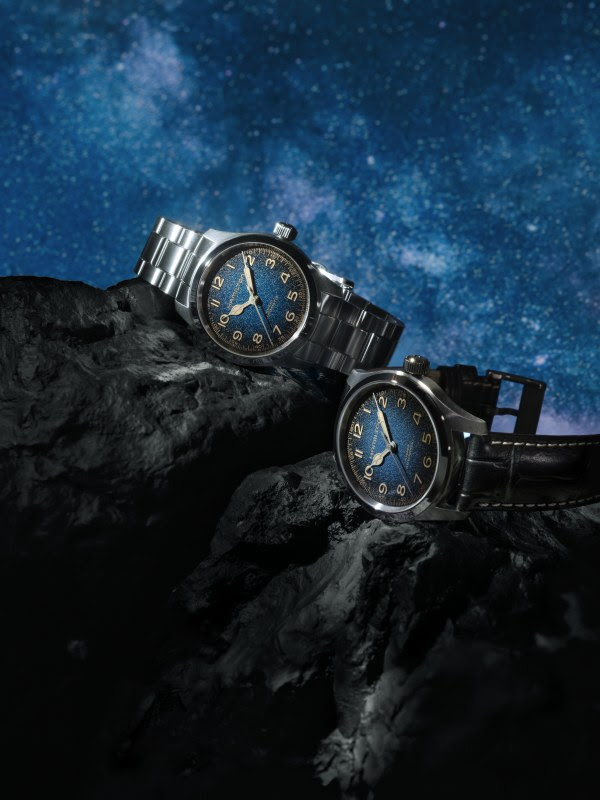 Two wristwatches with blue dials resting on a dark rocky surface beneath a starry night sky; one with a metal bracelet, the other with a leather strap.