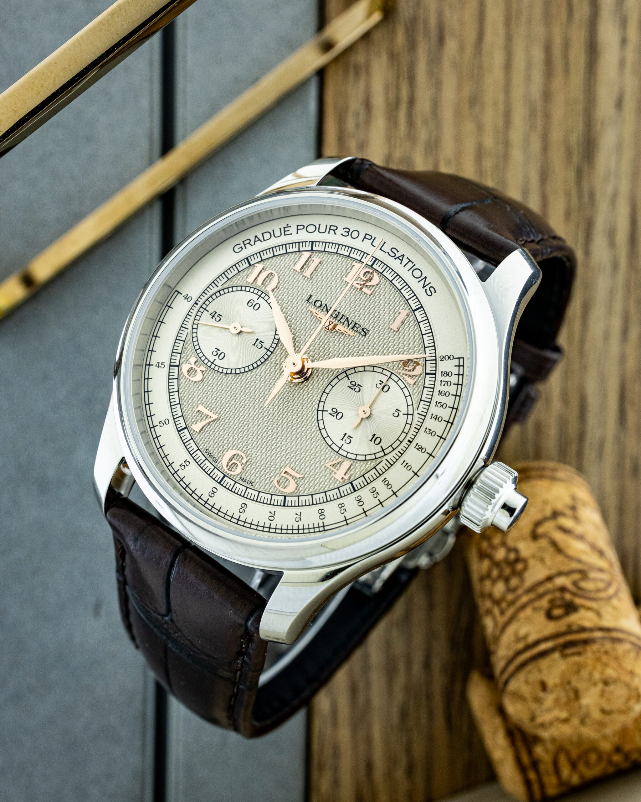 Close-up of a silver chronograph wristwatch with a textured beige dial and brown leather strap on a wooden surface.
