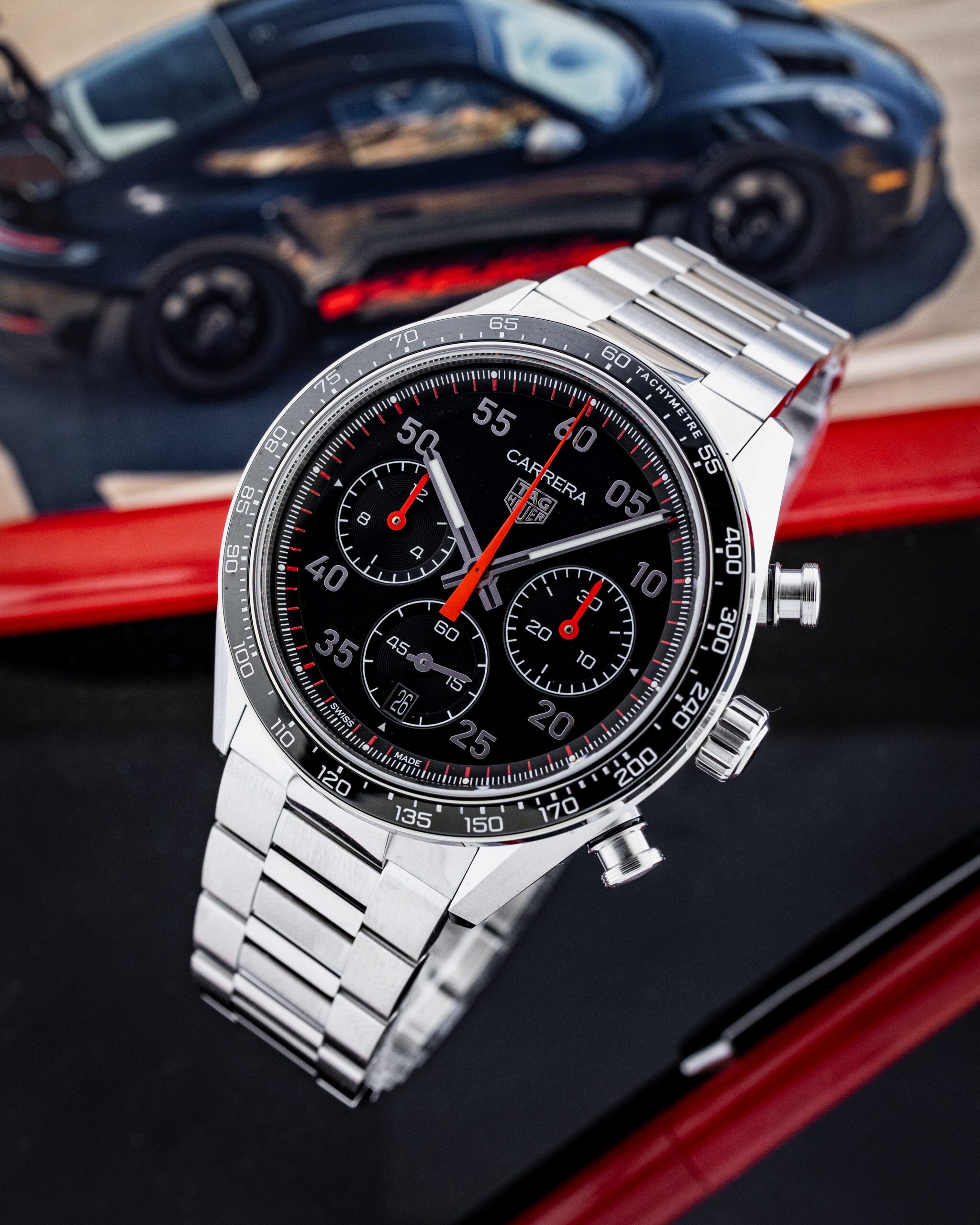 Close-up of a stainless steel chronograph watch with a black dial and red accents, tachymeter bezel, and multiple subdials, set against a blurred dark sports car background.