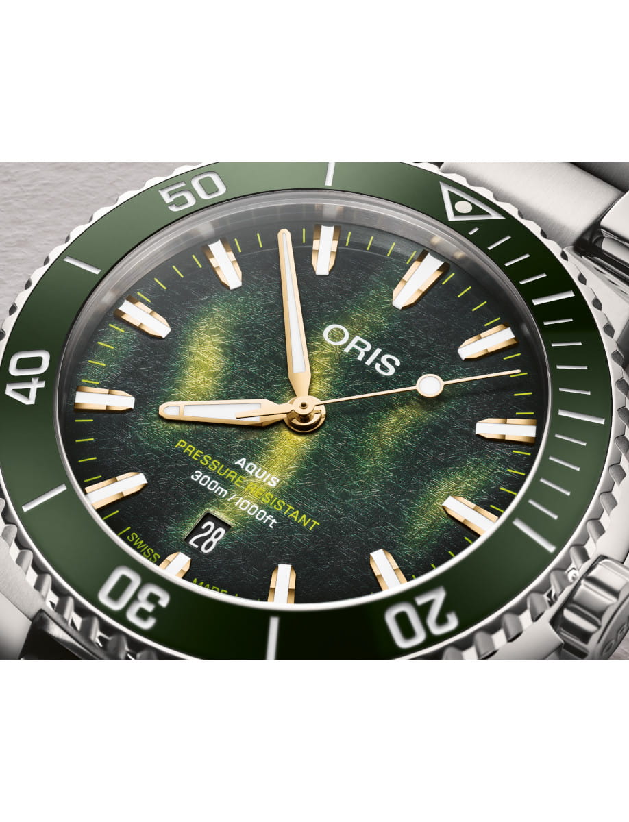 Close-up of a green-bezel Oris Aquis dive watch with gold hands and a textured green dial, showing the date at 6 o'clock and stainless steel case.