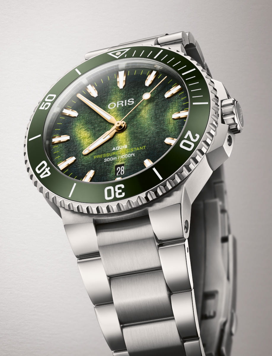 Close-up of a stainless steel ORIS Aquis dive watch with a green bezel, textured green dial, and date window at 3 o'clock on a stainless steel bracelet.