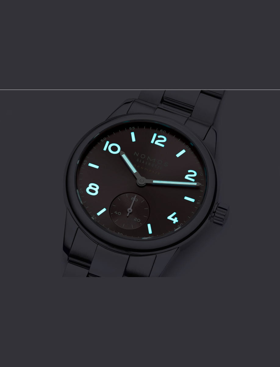 Nomos stainless steel wristwatch with glowing blue numerals and hands, tilted on a dark background, showing a small subdial at 6 o'clock and a prominent crown on the right side.