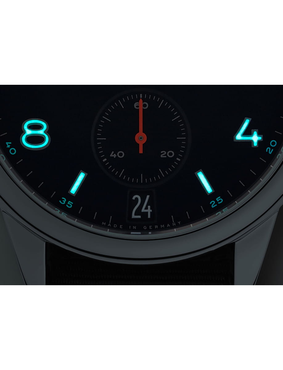 Close-up of a black watch dial with glowing blue markers at 8 and 4, a red central second hand, and a 24 date window; 'Made in Germany' at the bottom.