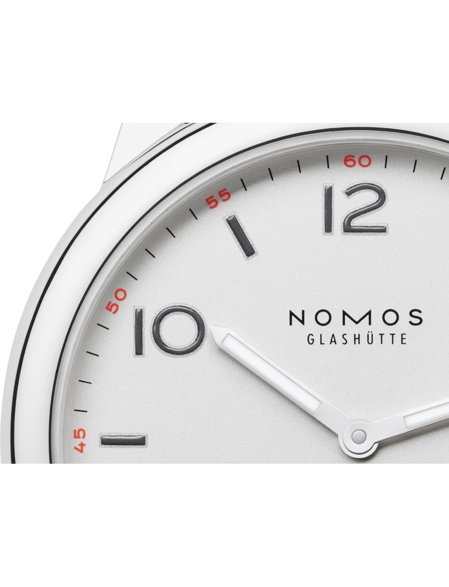 Close-up of a Nomos Glashütte watch dial with white hands and silver hour markers on a light face.