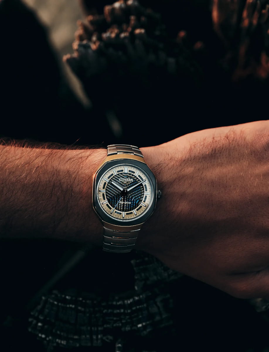 Wristwatch on a metal band with a blue and silver face, worn on the left wrist outdoors at dusk (Citizen watch).