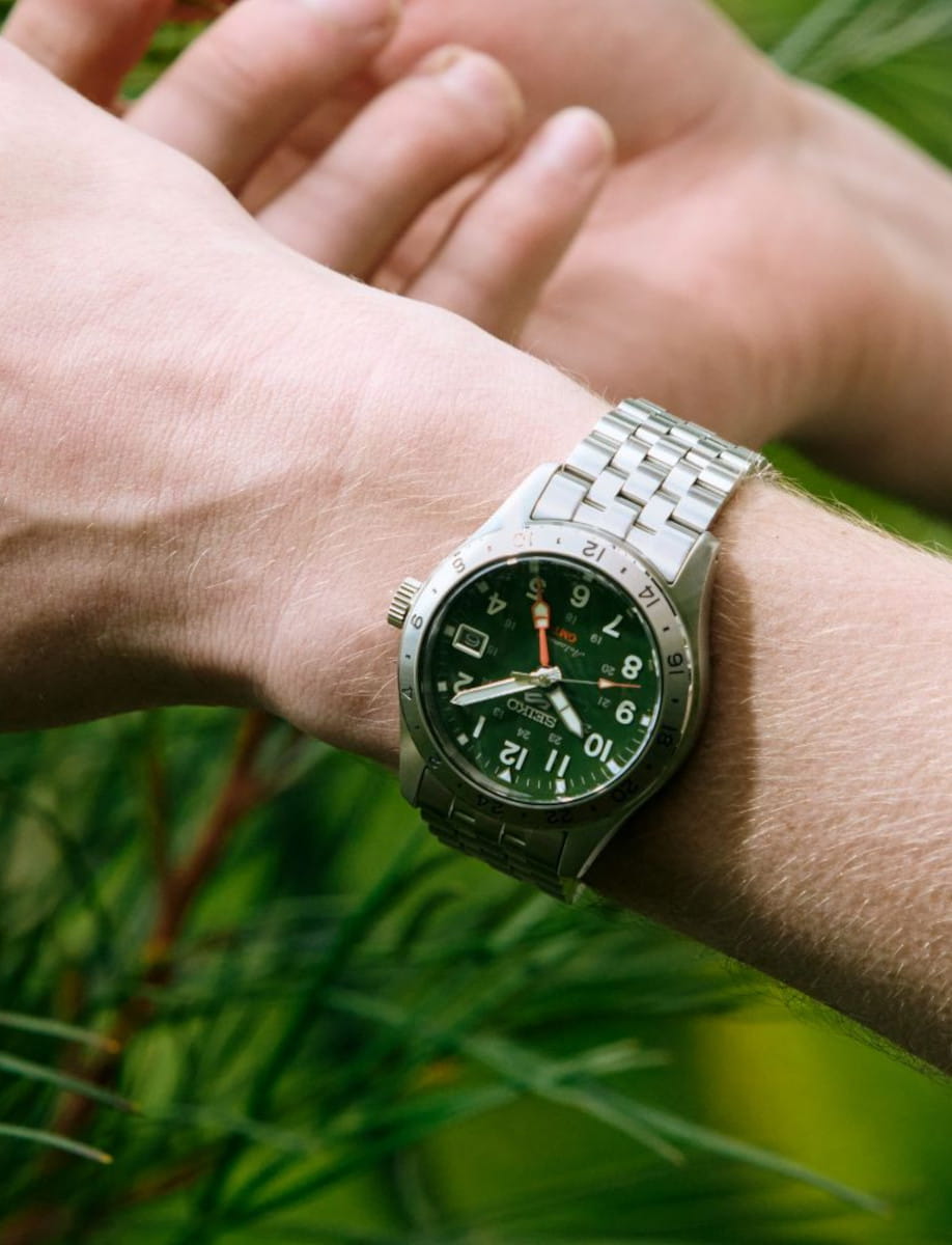 Seiko stainless‑steel wristwatch with a green dial on a forearm, outdoors among green foliage in the background