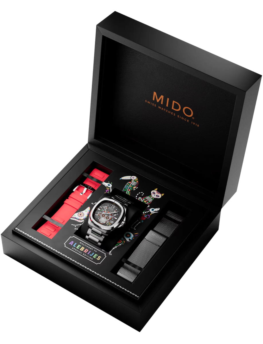 Open black MIDO presentation box showing a metal-cased wristwatch with multiple straps (red and metal) and a colorful insert.