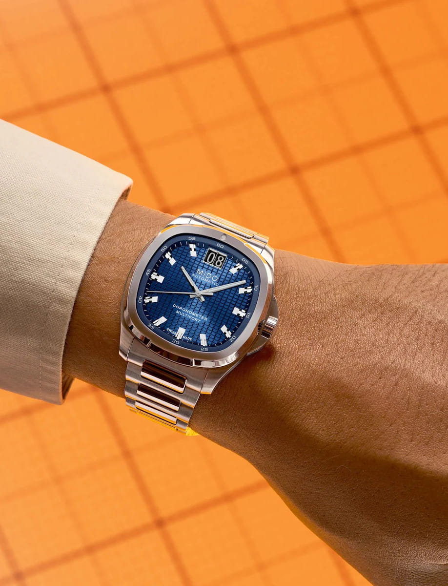 Close-up of a Mido stainless steel wristwatch with a blue textured dial on a person's wrist against an orange background.
