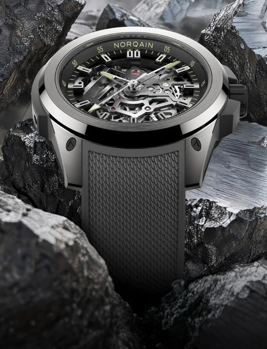 Skeleton dial watch with exposed gears, black bezel reading NORQAIN, metal case and textured rubber strap among rocks for rugged, high-end appeal.