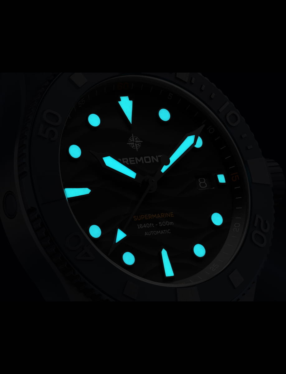 Close-up of a Bremont watch dial with bright blue luminous on hour markers, hands, and bezel numbers, showing a glowing timepiece in low light.