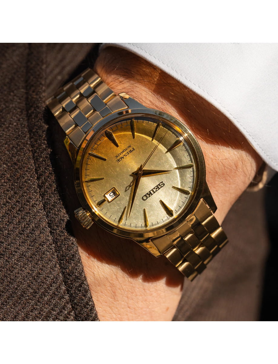 Gold-tone Seiko automatic wristwatch worn on the left wrist, with a gold tone steel bracelet and a white shirt cuff nearby.