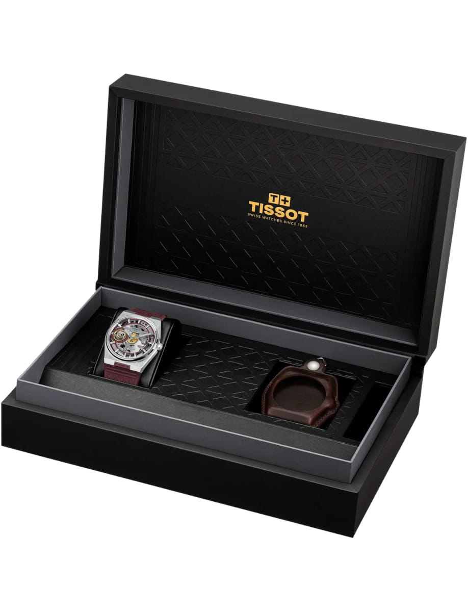 Open black watch box displaying a silver skeleton watch with burgundy strap in the left slot and an additional leather strap.