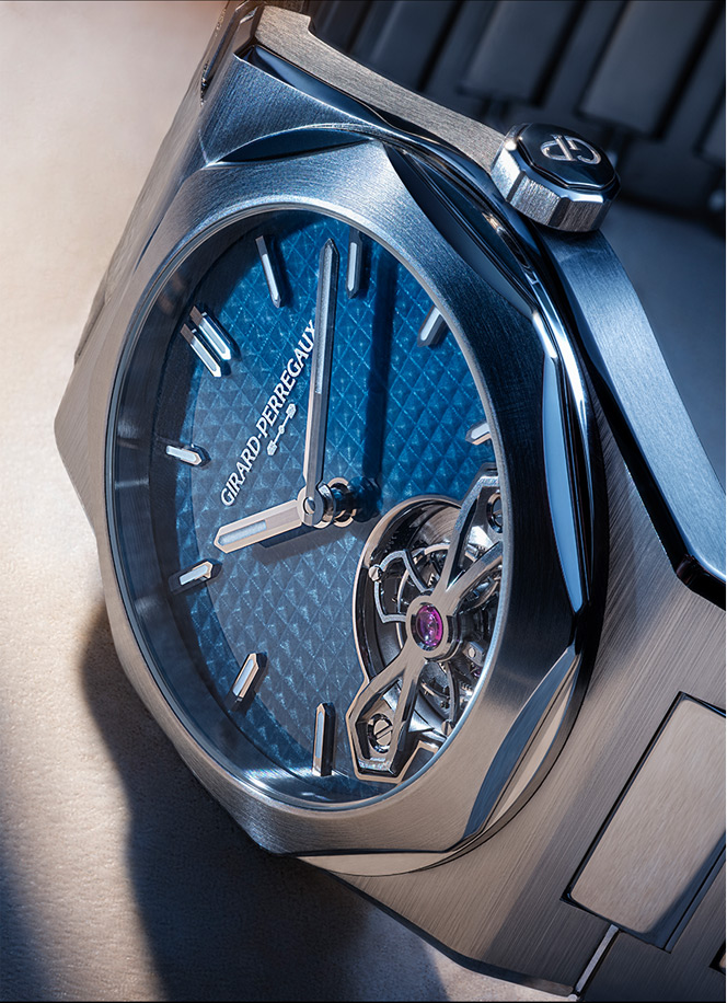 Close-up of a blue Girard-Perregaux watch with a visible tourbillon and textured dial, stainless-steel case visible from the side.